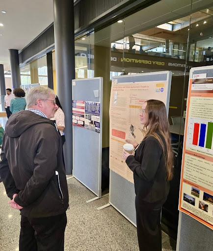 A woman with long hair and glasses explains her urban agriculture poster to an attendee.