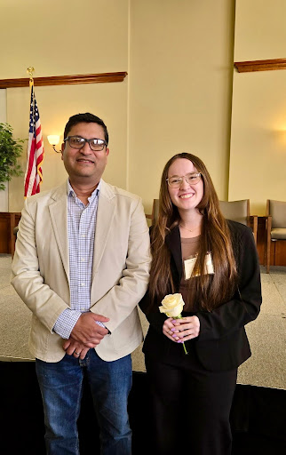 Meenar in a tan blazer and student Nicole in a black suit stand in front of a stage. Nicole is holding a white rose.